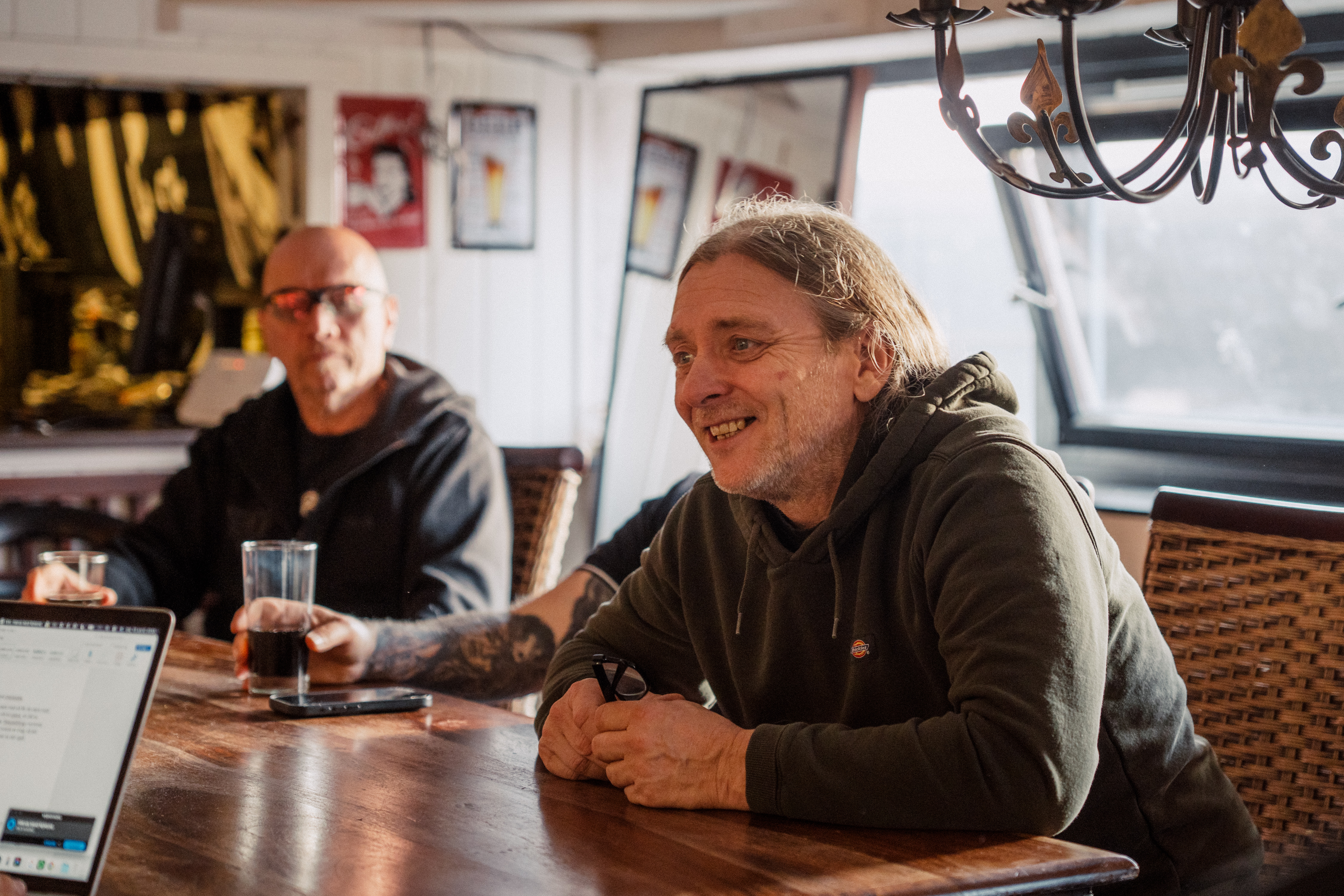 Tre menn sitter rundt et trebord i et møterom. Stein-Roger Arnøy sitter og smiler i forgrunnen med grønn hettegenser, mens de to andre deltar i samtalen.